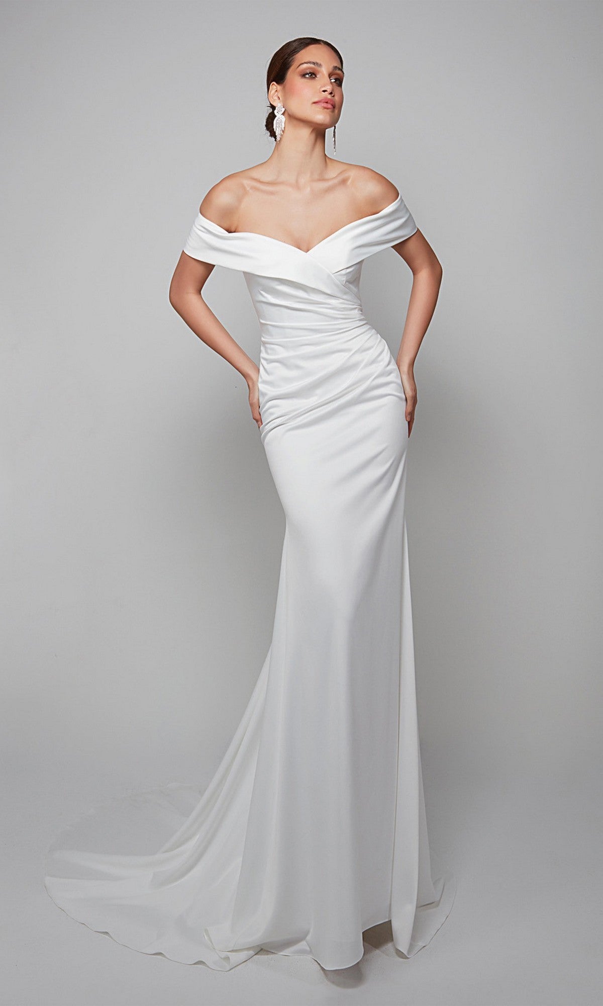 A woman stands against a plain light background wearing the Long White Dress: Alyce 7059 in Diamond White, a fitted satin formal gown featuring an off-shoulder design and a slight train.